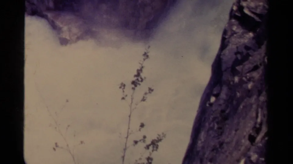 1979 Canada 8mm frame with pale waterfall mist, rushing water, a dark film border, sparse branches, and rocky forms near the gorge edge.