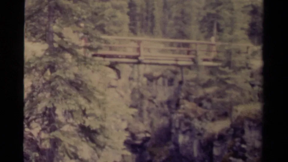 Faded 1979 8mm Canada frame of a wooden pedestrian bridge spanning a steep evergreen ravine.