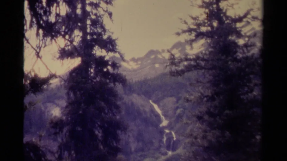 1979 Canadian 8mm mountain view with evergreen branches, distant slopes, snow patches, and a narrow bright waterfall or stream.