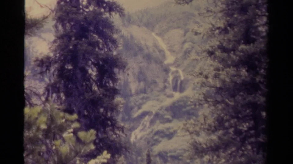 1979 Canadian home-movie frame with trees in the foreground and a hazy mountain valley crossed by a bright watercourse.