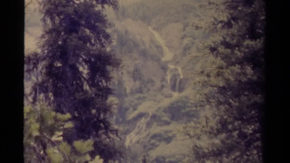 Final 1979 Canadian 8mm frame showing foreground evergreens, soft mountain slopes, and a distant bright stream or waterfall.