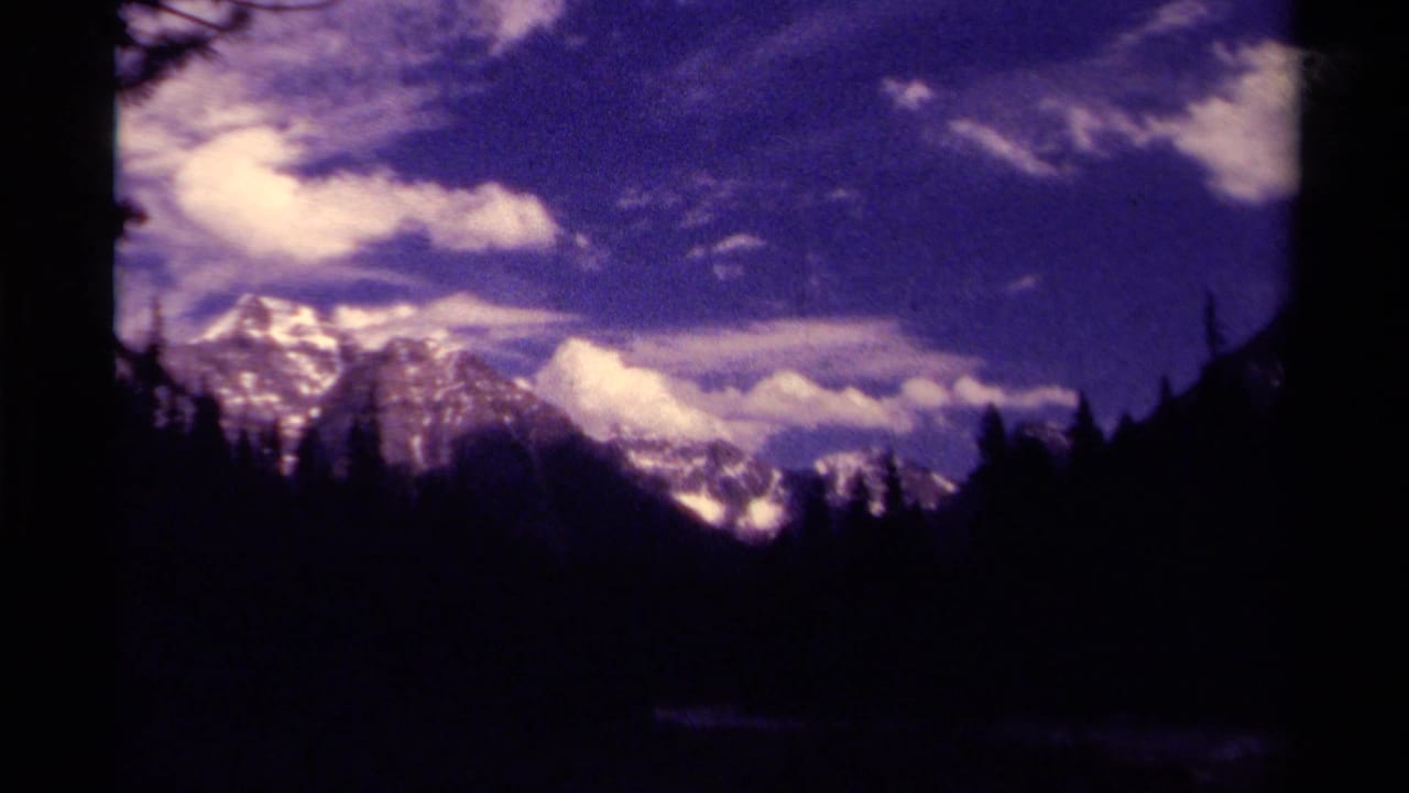 1979 8mm home-movie view in Canada of snow-capped mountains, dark trees, and a cloud-filled sky with purple film cast.