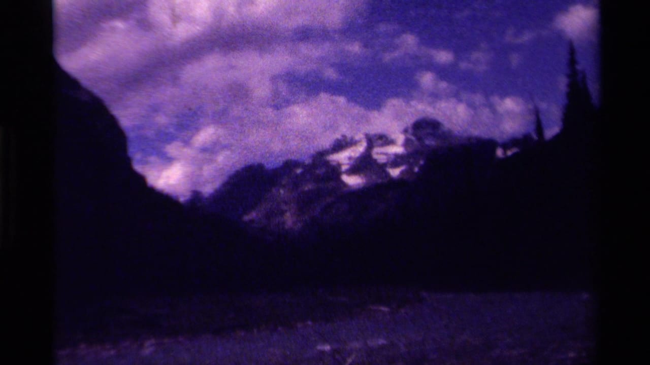 Canadian mountain landscape on 8mm film, showing a dark valley, snow-bright peaks, and dense cloud forms in purple-tinted footage.