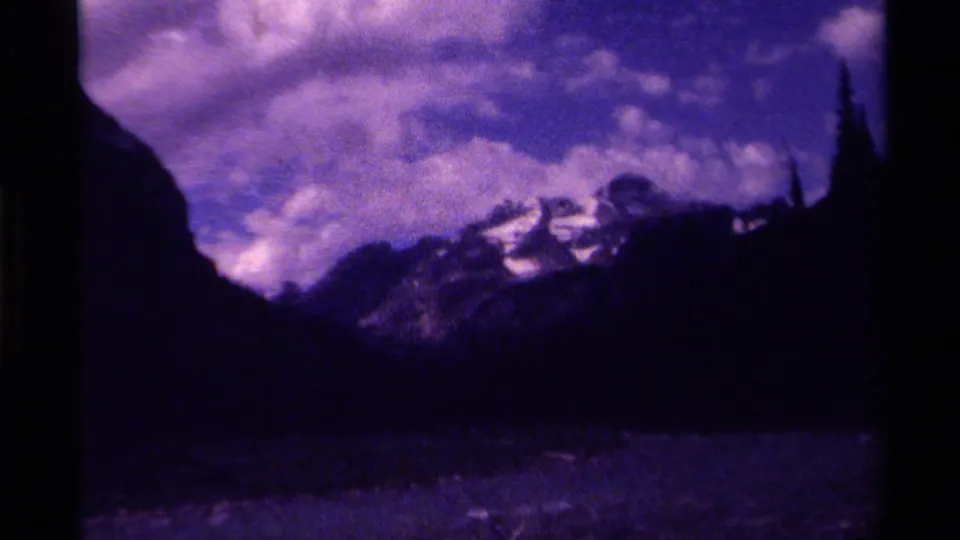 Canadian mountain landscape on 8mm film, showing a dark valley, snow-bright peaks, and dense cloud forms in purple-tinted footage.
