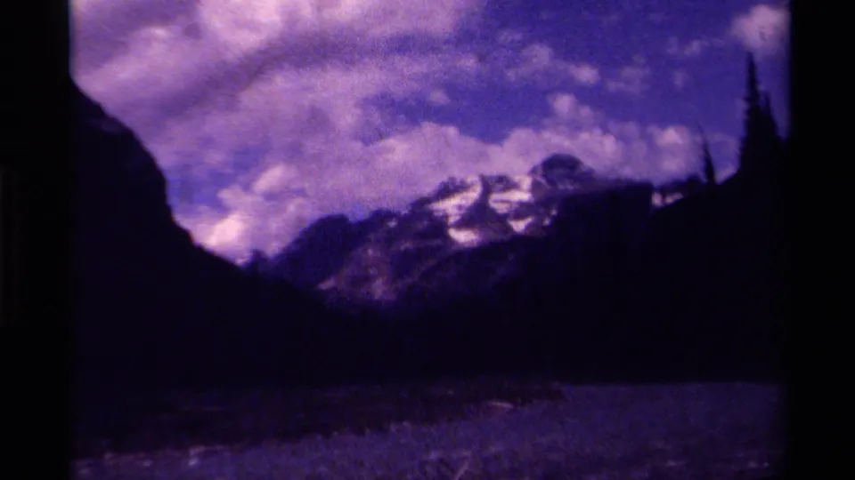 Purple-cast 8mm home-movie still from Canada showing a snowy mountain ridge, dark valley, and heavy cloud cover.