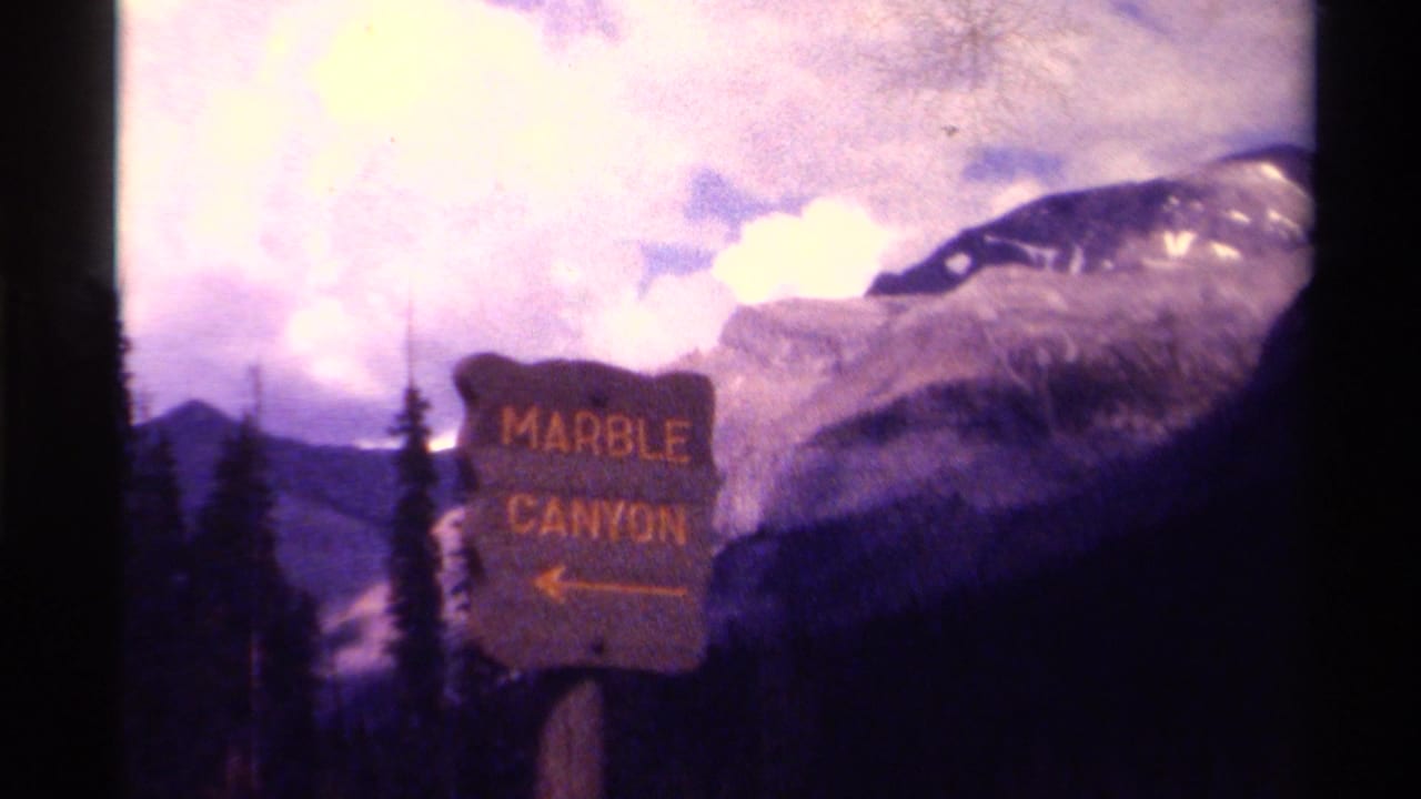 Marble Canyon sign beside evergreen trees and mountains under a cloudy sky.