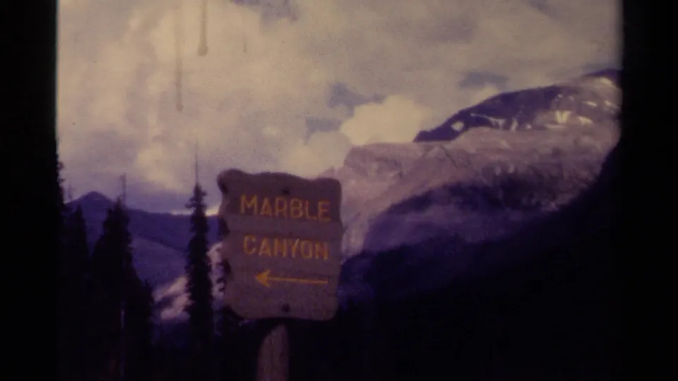 Canada 1979 8mm frame of the Marble Canyon wayfinding sign set before conifers, mountain slopes, snow patches, and a cloudy sky.