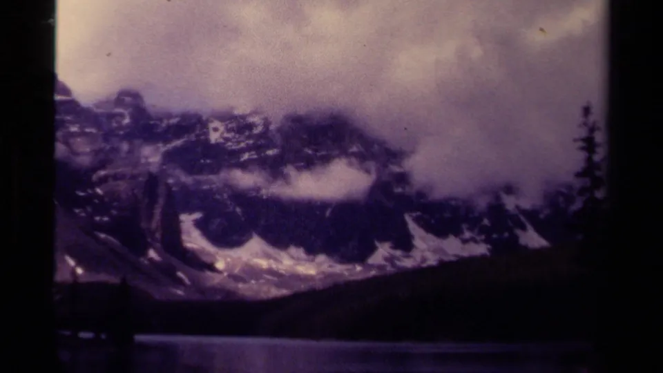 1979 Canadian 8mm frame showing snow-capped mountains, cloud, a lake, and a dark forested shore.
