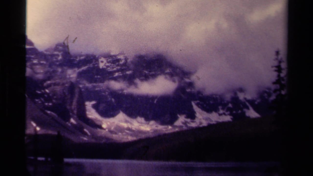 Misty snow-streaked mountain range above a dark lake with evergreen silhouettes.