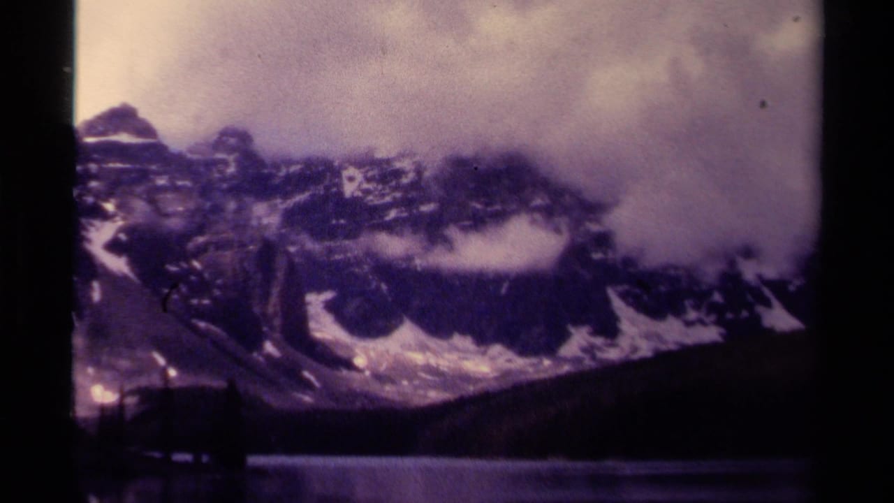 Canadian mountain range appears under low cloud, with snow patches, dark forest, and lake foreground in a 1979 8mm frame.