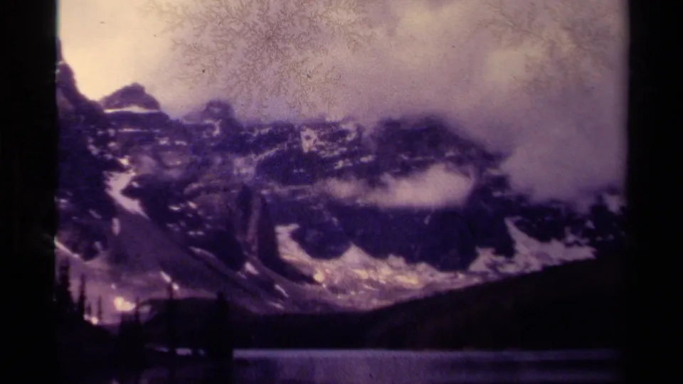 Soft 8mm frame of Canadian mountains partly hidden by cloud, with a dark lake, tree line, and visible film texture.