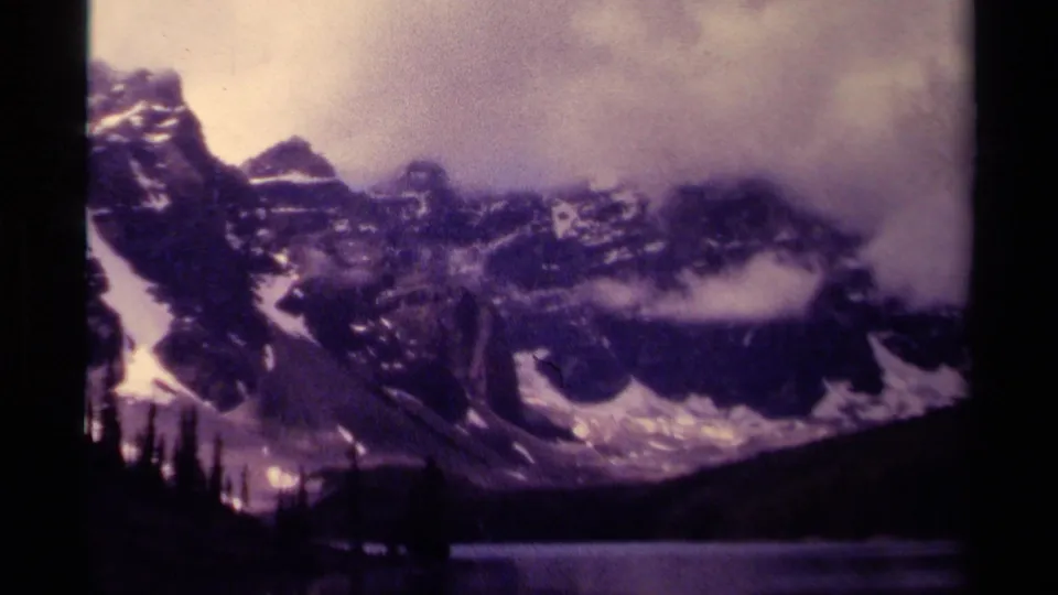 1979 Canadian home-movie frame with mist covering a snowy mountain face above a dark lake and evergreen shoreline.