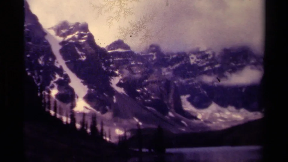 1979 Canadian home-movie frame of rugged snow-covered mountains under low cloud with evergreens and lake shoreline below.