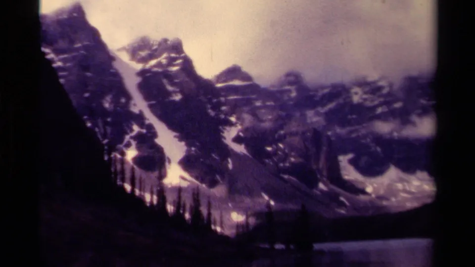 1979 Canadian 8mm frame with a closer view of snow-lined mountain slopes, evergreen silhouettes, cloudy sky, and lake water near the bottom.