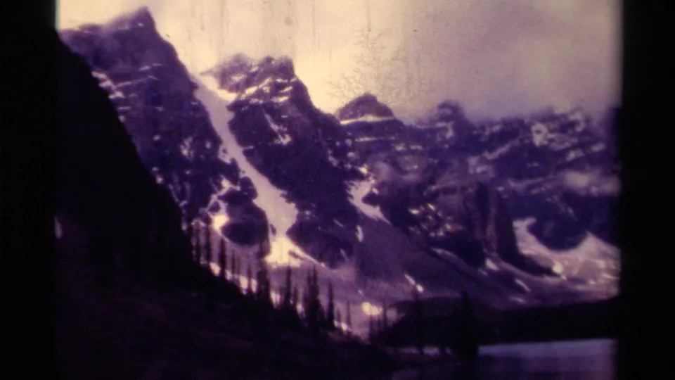 1979 Canadian 8mm frame showing snow-streaked peaks, a dark evergreen shoreline, cloudy sky, and lake edge within rounded film borders.