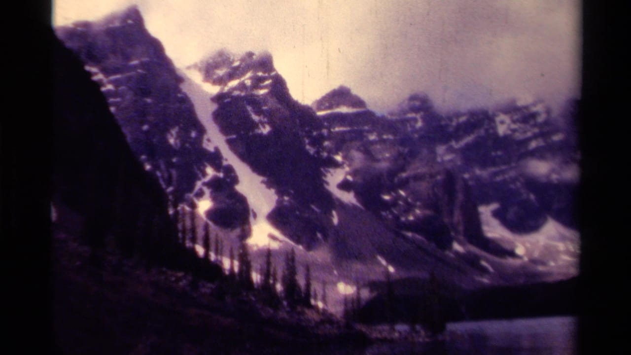 Snow-capped mountain peaks above a dark lake with conifer silhouettes in the foreground.