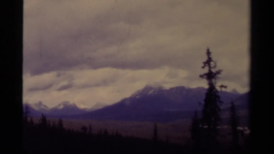 1979 Canada 8mm landscape frame shows distant snow-capped mountains, evergreen silhouettes, dark foreground trees, and a cloudy sky.