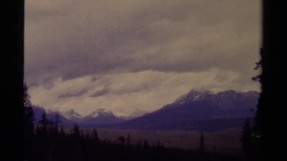 A distant Canadian mountain range appears through 1979 8mm color fade, with a low forest line and heavy cloud cover.