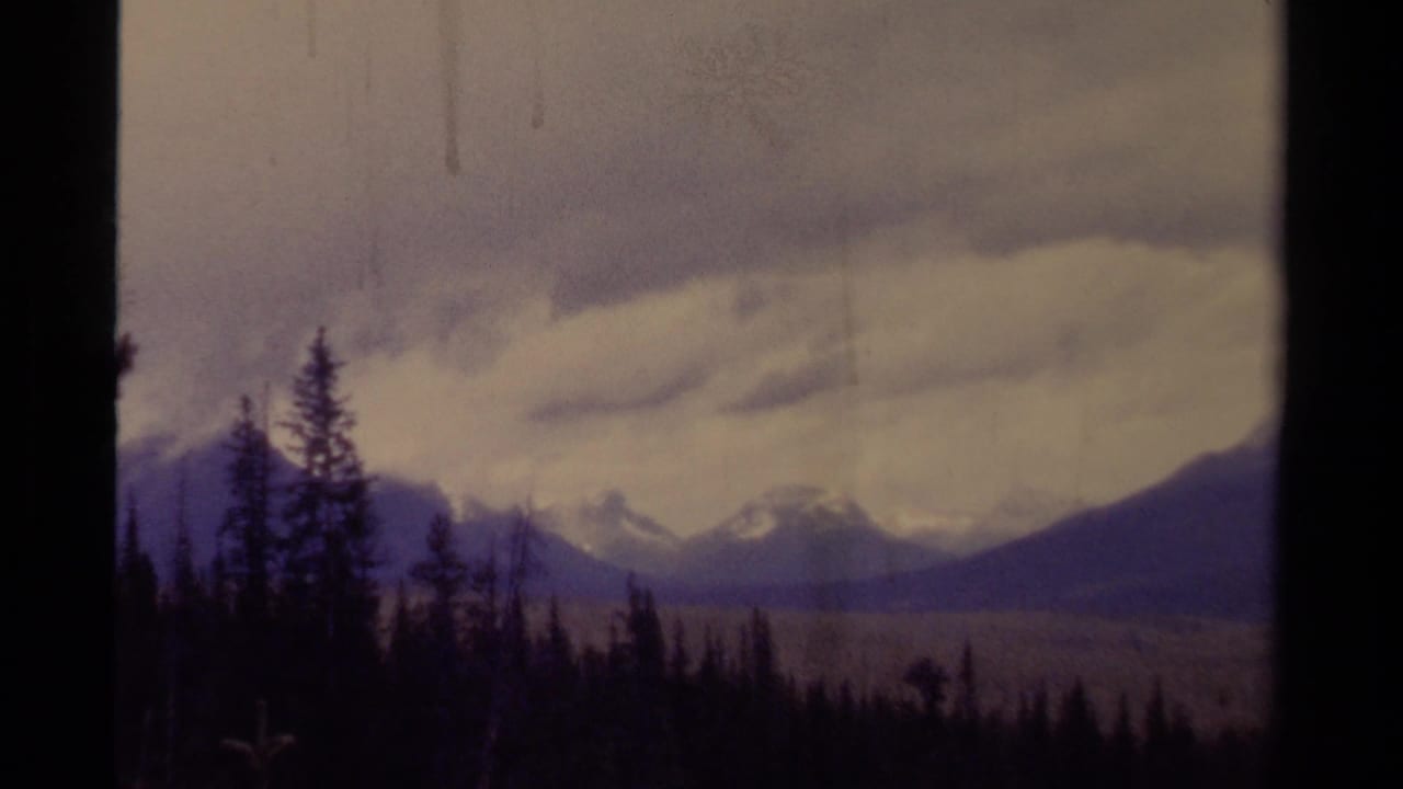 1979 8mm Canada frame showing dark evergreen forest, distant mountain peaks, and a heavy overcast sky.