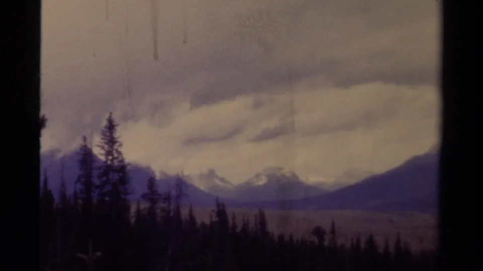 1979 8mm Canada frame showing dark evergreen forest, distant mountain peaks, and a heavy overcast sky.