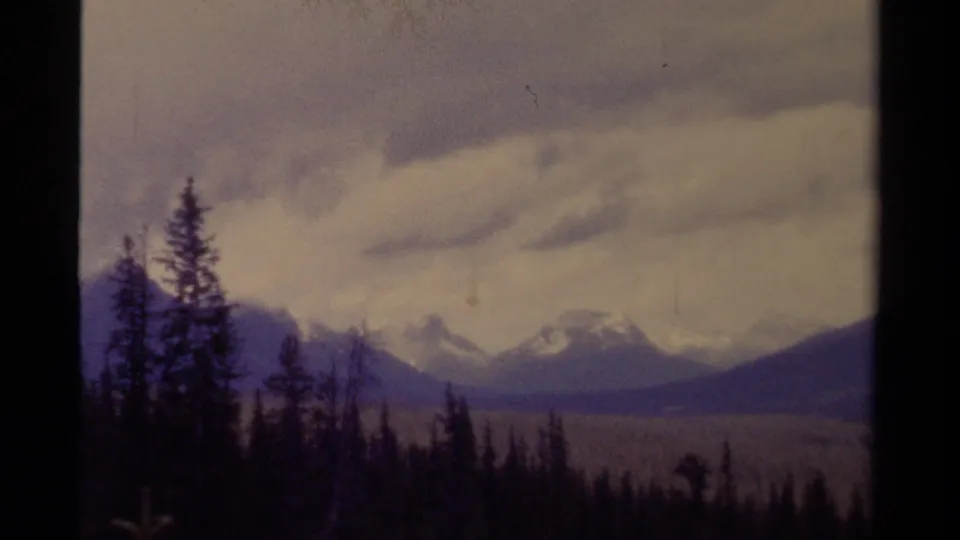 1979 8mm Canada view of evergreen silhouettes and snow-dusted mountains under thick cloud cover.