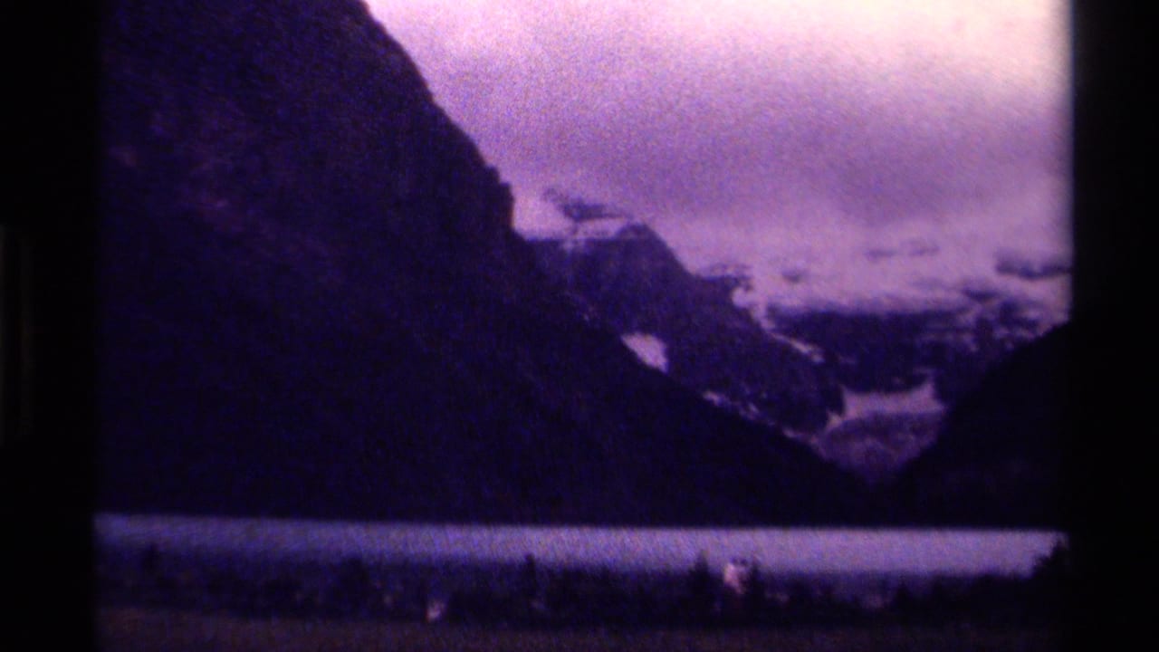 Canadian 8mm landscape frame showing a mountain lake, dark evergreen foreground, steep slopes, snowfields, and purple film cast.