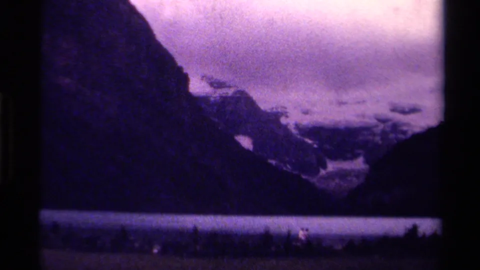 1979 Canadian wilderness view on 8mm film, with a dark mountain slope, lake band, evergreen shoreline, and distant snow-capped peaks.