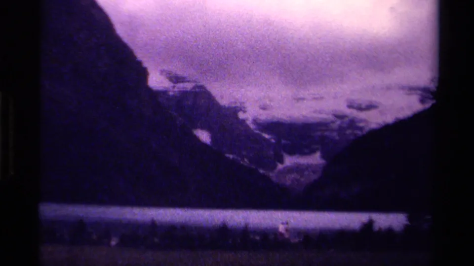 Canadian 8mm home-movie frame of a cloudy mountain lake, dark evergreen foreground, steep slopes, and distant snowfields.