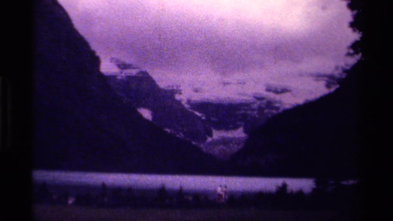 1979 8mm Canada home-movie frame showing a mountain lake beneath cloudy peaks, with small people near the shoreline.