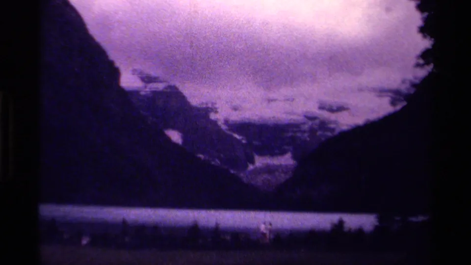 1979 8mm Canada home-movie frame showing a mountain lake beneath cloudy peaks, with small people near the shoreline.