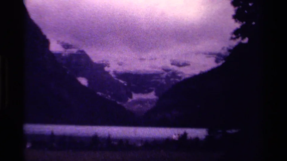 Canadian 1979 home-movie frame showing a purple-toned mountain lake, dark tree silhouettes, and small shoreline figures.