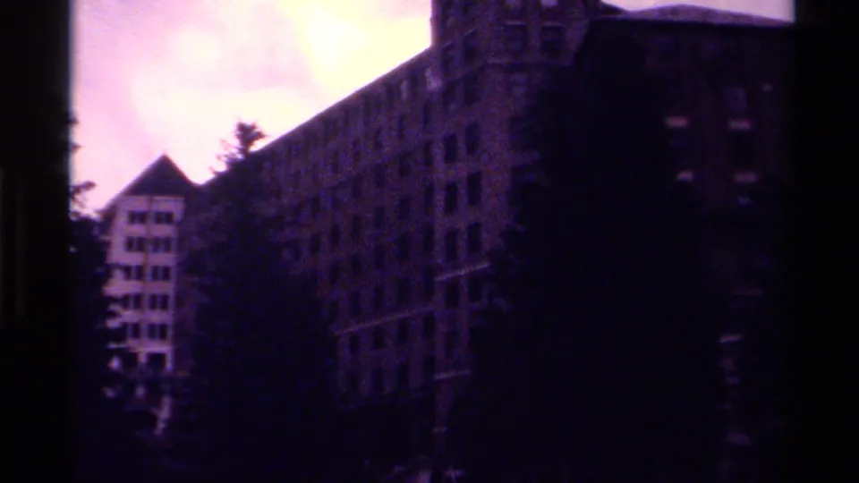 1979 Canada home-movie frame of a large multi-story building with many windows, partly obscured by tall trees.
