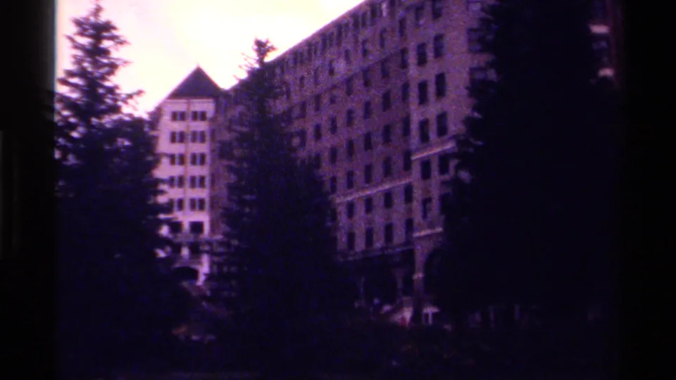 Final 1979 Canada home-movie frame with a large windowed building framed by pine trees and a dark foreground near water or shoreline.