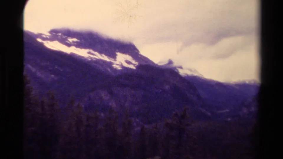 Canadian mountain vista on 8mm film, with snow patches, forested slopes, and low clouds.