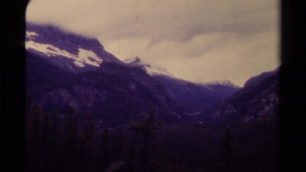 Mountain valley with snow patches, steep slopes, and a cloudy pale sky.