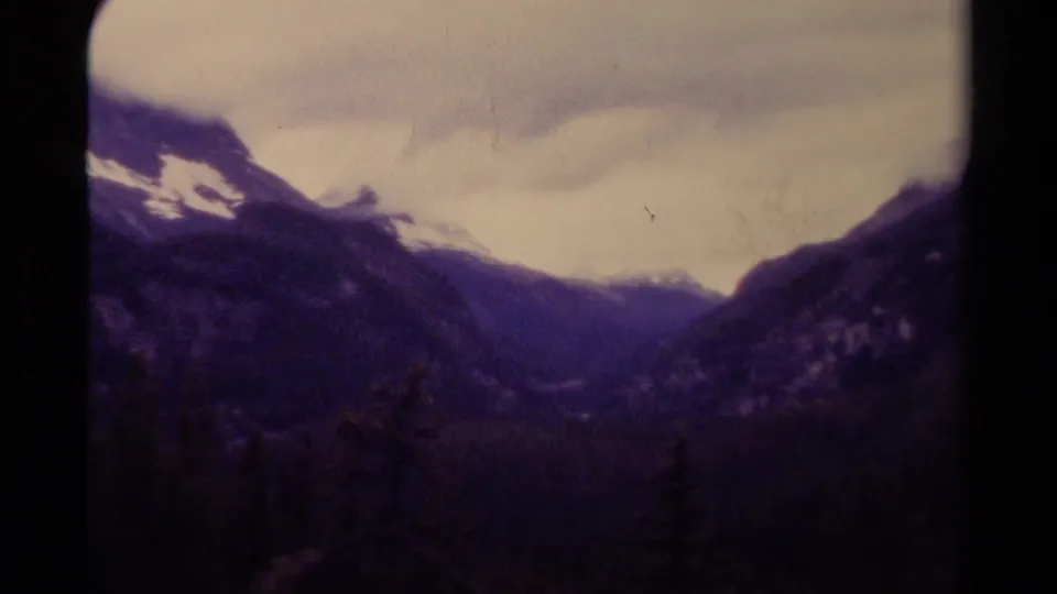 1979 Canadian 8mm frame with a shadowed mountain valley, snow along the left ridge, and dense pale clouds.