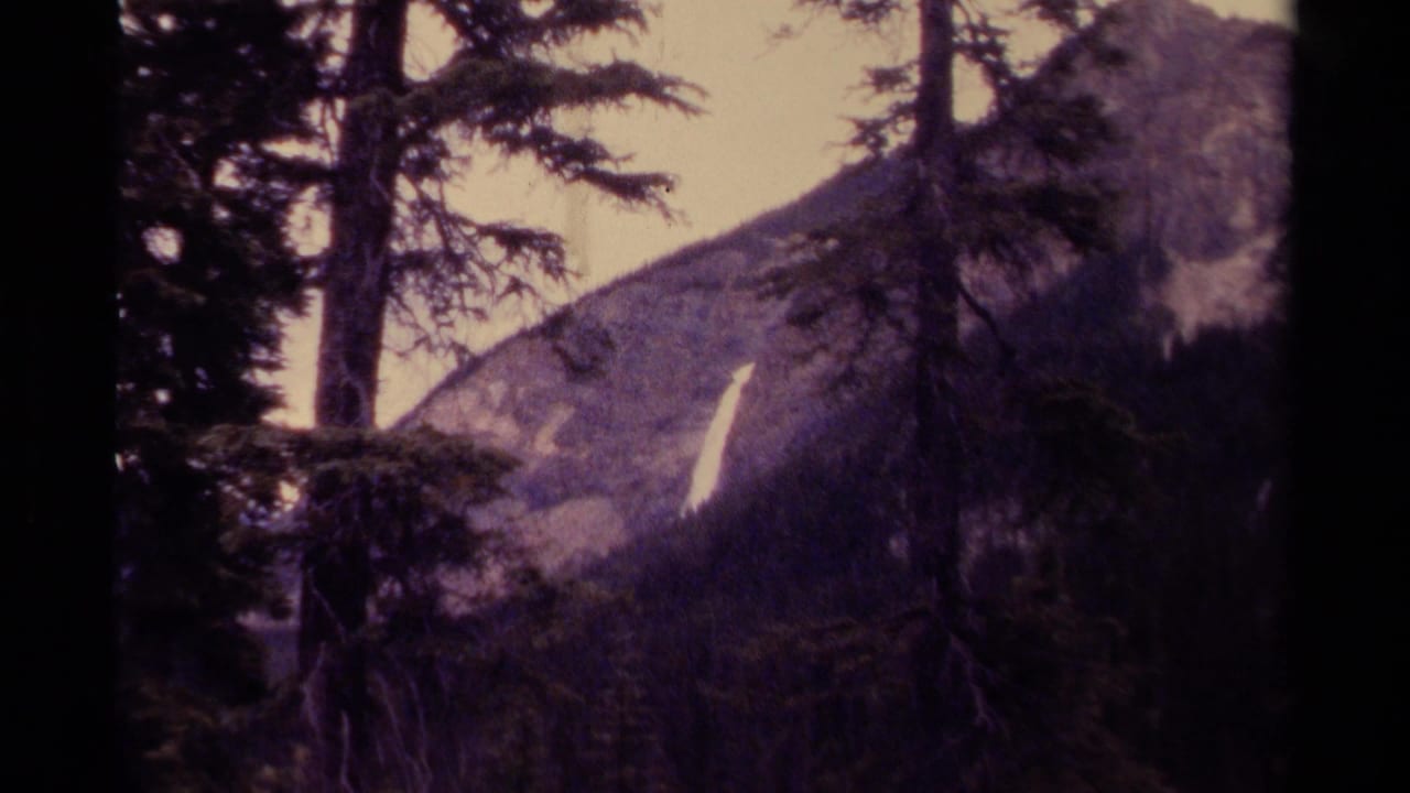 1979 Canadian mountain view with foreground evergreens and a slim waterfall cutting down a rocky slope in soft 8mm color.
