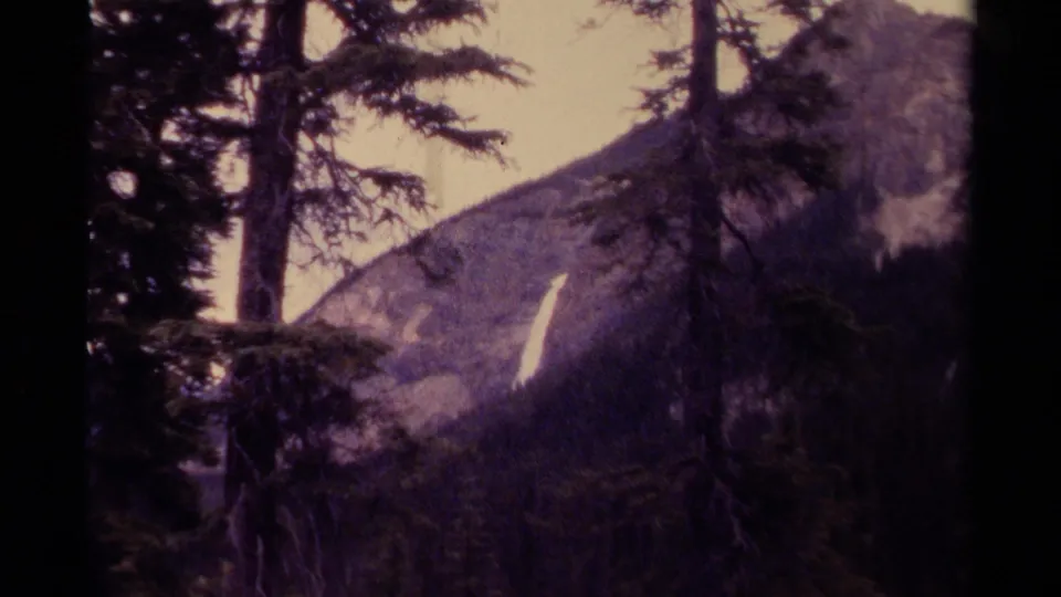 1979 Canadian mountain view with foreground evergreens and a slim waterfall cutting down a rocky slope in soft 8mm color.