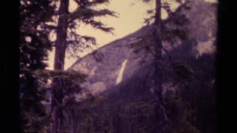 A handheld 8mm view in Canada shows evergreens in the foreground and a bright waterfall descending a mountain slope.