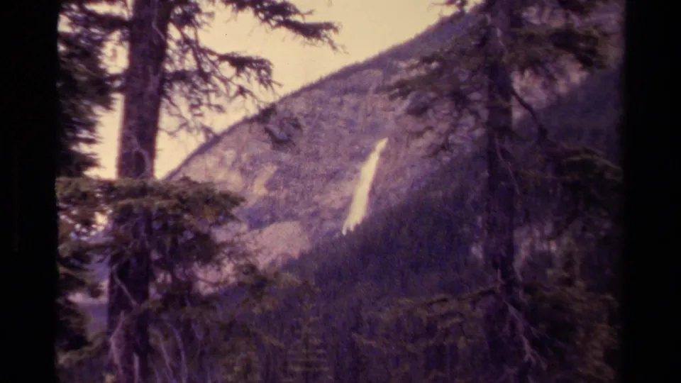 1979 Canada home-movie frame with purple-tinted grain, evergreen silhouettes, rocky slope, and a narrow white waterfall.