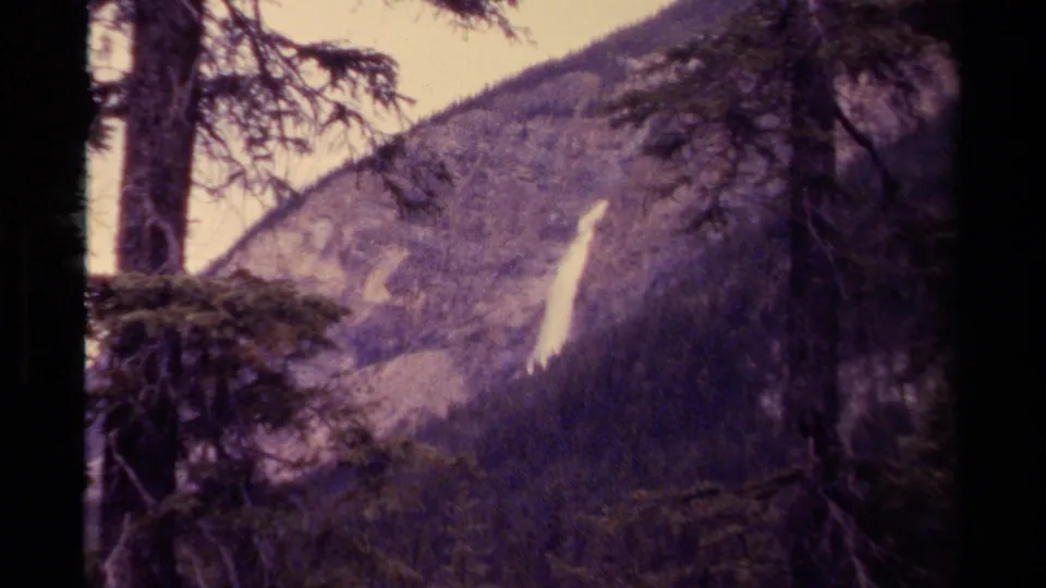 1979 Canada 8mm frame showing a bright waterfall on a wooded hillside, viewed through dark evergreen trees.