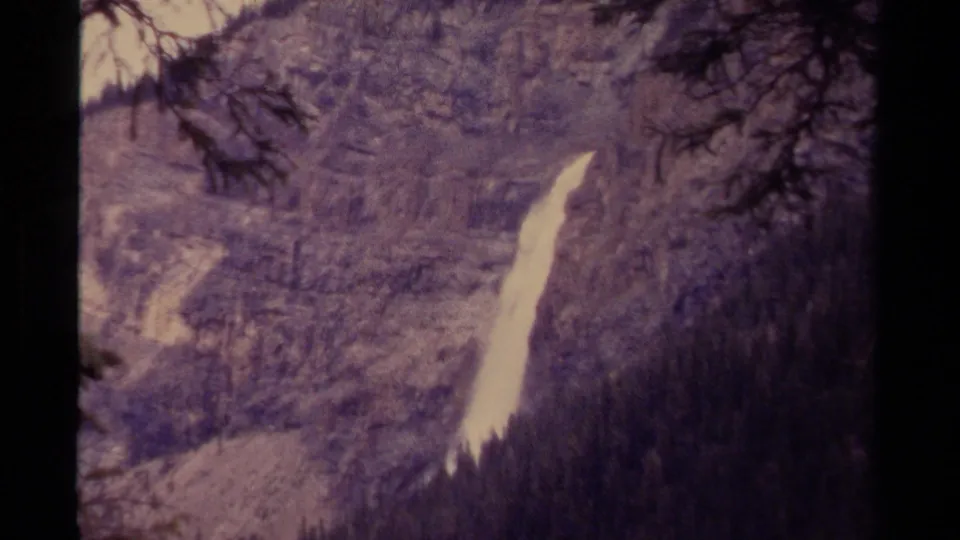 Faded 8mm frame of a Canadian waterfall on a forested slope, partially obscured by nearby trees.