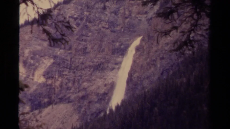 1979 Canadian 8mm frame showing a distant waterfall descending a rocky cliff, partially framed by dark trees and valley forest.