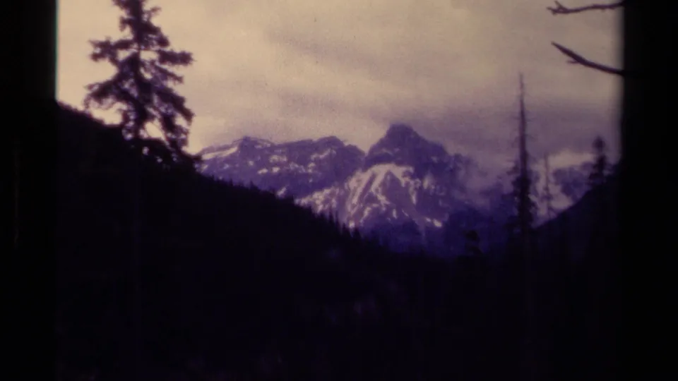 1979 Canadian 8mm frame of distant snow-streaked mountain peaks seen from a shadowed forest valley under cloud cover.