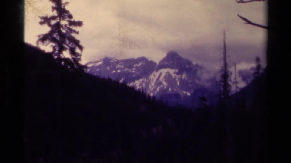 1979 Canadian 8mm frame with a distant ridge and pointed peak beyond silhouetted evergreens in a shaded valley.