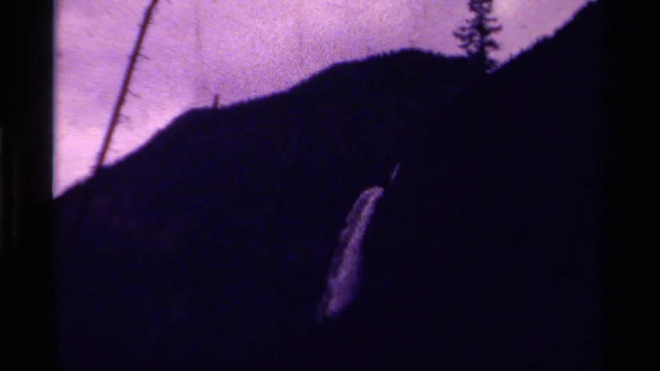 Grainy 1979 8mm view of a bright waterfall descending a shadowed Canadian mountain slope beneath a purple sky.