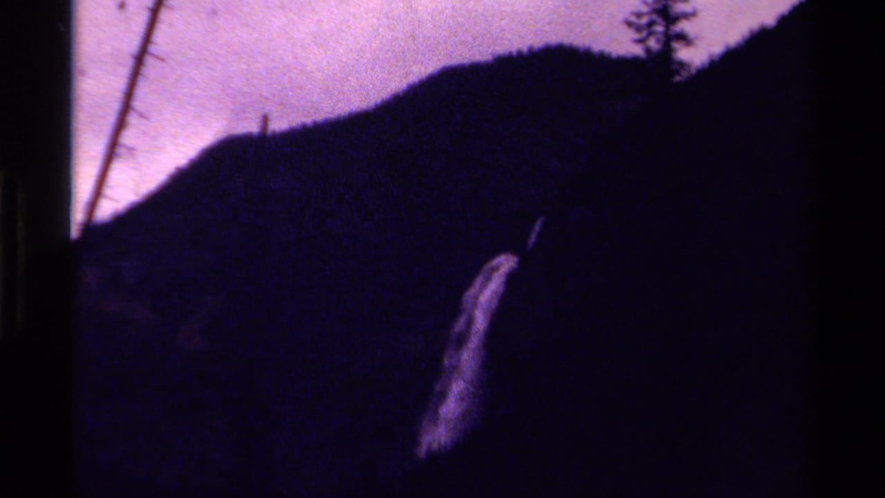 1979 Canadian 8mm frame showing a distant waterfall dropping along a shadowed mountain slope with trees and purple color shift.