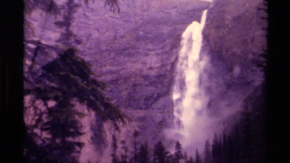 Closer 1979 Canadian 8mm frame of a tall waterfall pouring down a forested slope, with pine branches in the foreground and mist below.