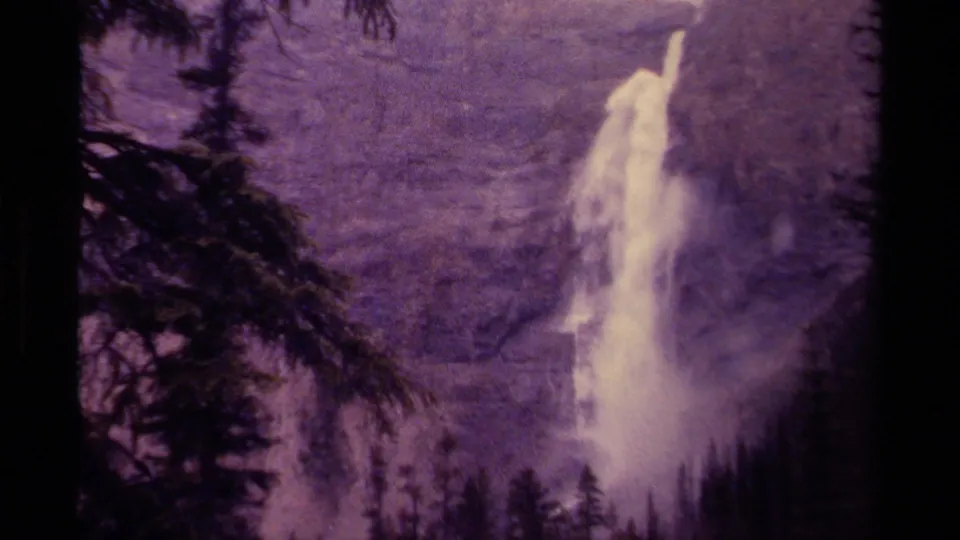 1979 Canadian 8mm frame showing a tall white waterfall cascading down a rugged cliff beside dark evergreen forest.