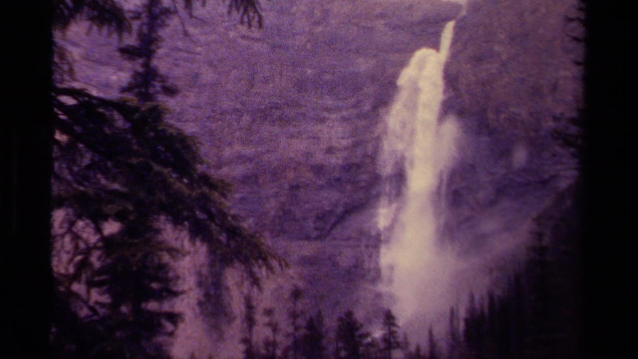 Distant white waterfall drops down a dark mountain face behind evergreen silhouettes.